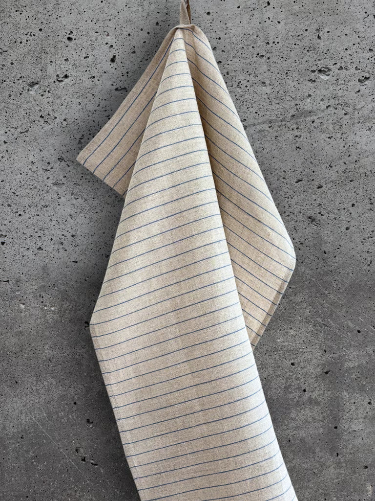Beige kjøkkenhåndkle i 100 % lin med brede blå striper, henger på en betongvegg.