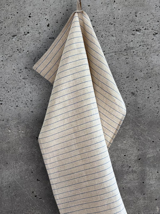 Beige kjøkkenhåndkle i 100 % lin med brede blå striper, henger på en betongvegg.