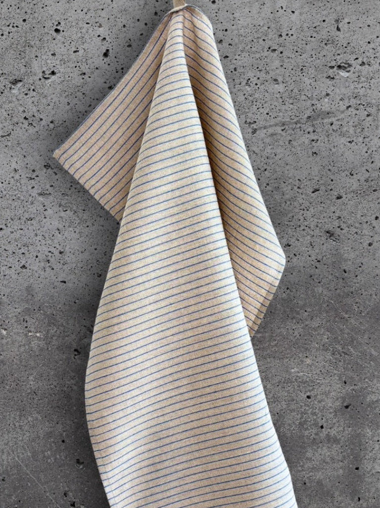 Stripete kjøkkenhåndkle i 100 % lin i beige med blå striper, henger på en betongvegg.