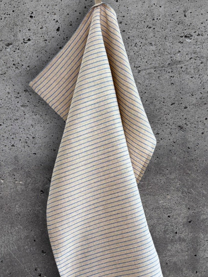 Stripete kjøkkenhåndkle i 100 % lin i beige med blå striper, henger på en betongvegg.