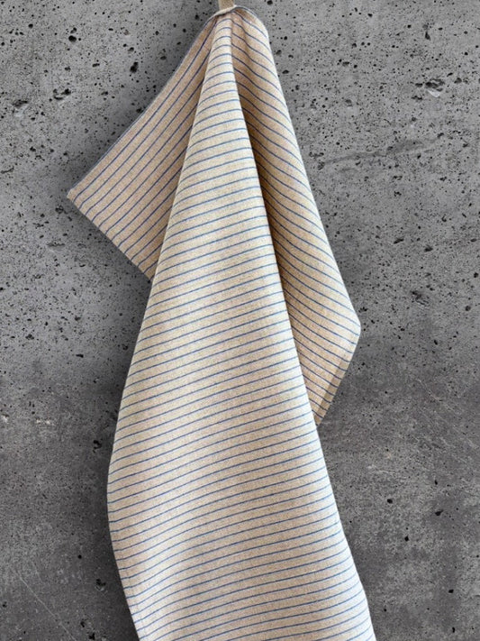 Stripete kjøkkenhåndkle i 100 % lin i beige med blå striper, henger på en betongvegg.